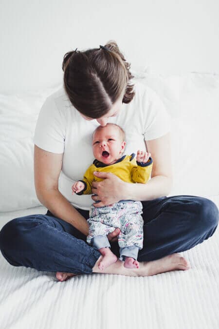 Mutter sitzt im Schneidersitz und küsst ihr Neugeborenes auf den Kopf welches sie vor sich hält