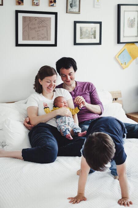 Junge tobt auf dem Bett herum. Im Hintergrund sitzten die Eltern mit einem Neugeborenen im Arm.