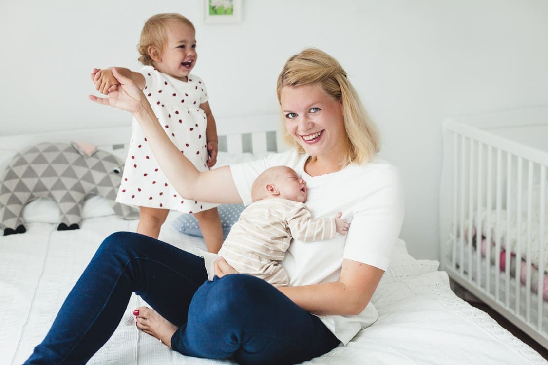Mutter sitzt mit ihren beiden kleinen Kindern auf dem Bett. Im Arm hält sie ihr Neugeborenes und an der Hand hält sie ihre kleine Tochter.