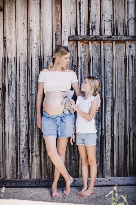 frau mit Babybauch und ihrer Tochter lehnen an einem Scheunentor. Die Tochter hat ein Kuscheltier in der Hand.