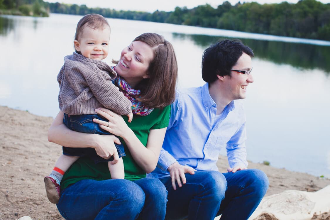Eltern mit kleinem Kind sitzen am See auf einem Baumstamm