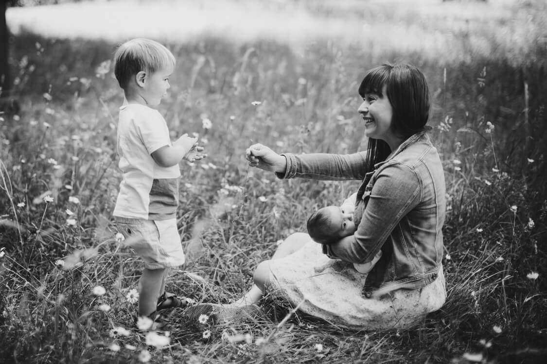 Mutter sitzt mit Baby auf einer Wiese. Der große Sohn pflückt der Mutter eine Blume.