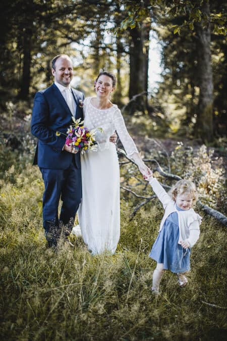 Brautpaar steht an einer Waldlichtung und Tochter zieht die Mutter am Arm