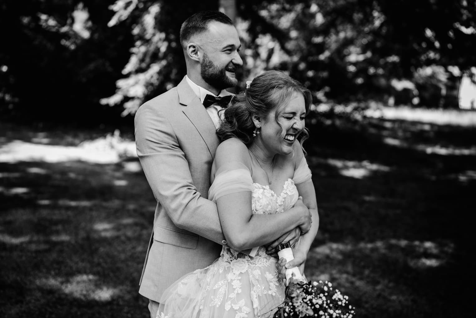 Schwarz-Weiß Hochzeitsfoto Bräutigam umarmt Braut von hinten und Braut lacht
