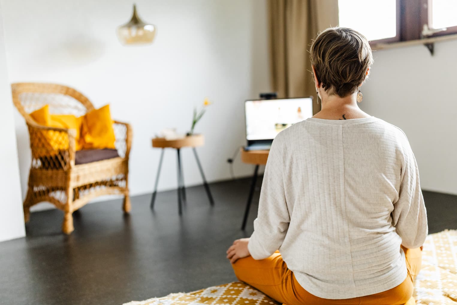 Frau von hinten im Schneidersitz mit Blick auf Laptop beim Online-Yoga-Kurs, entspannte Szene